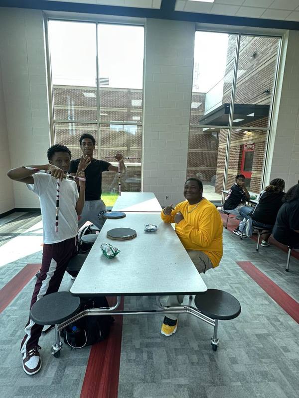 One student seated and two standing holing up fingers at a table in the cafeteria during the Perfect Attendance Celebration