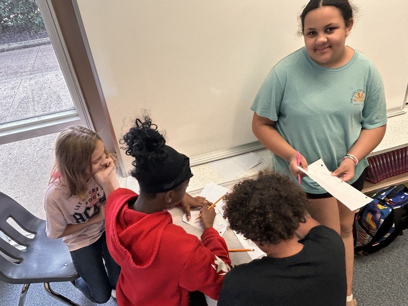 Students are working in a small group around a desk calculating unit rates after "grocery shopping" in the media center.