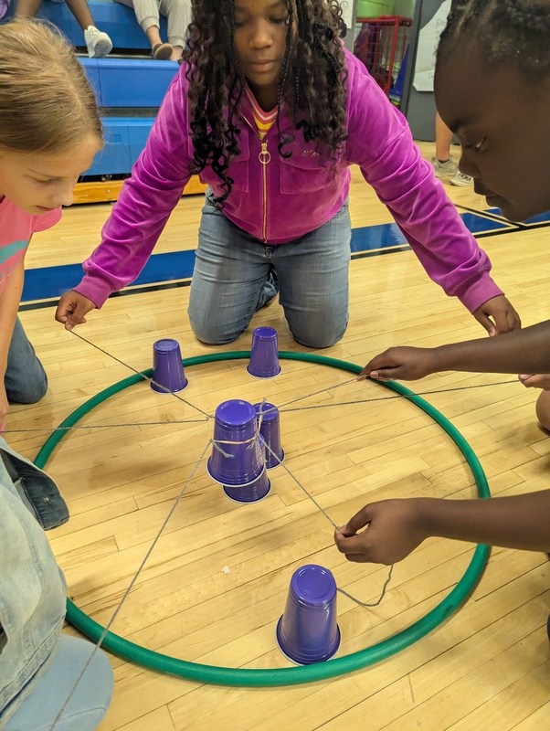 Students are working on the gym floor during Physical Education class working in a small group to stack cups using strings to grab, move and stack the cups. This is a team building exercise that required communication and teamwork.