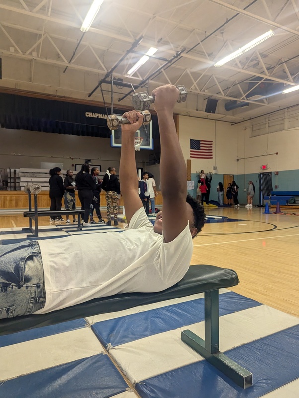 A student is lying on the bench using free-weights during Physical Education class in the gym. This was a unit study focusing on basic weight-lifting techniques and fitness goal setting.