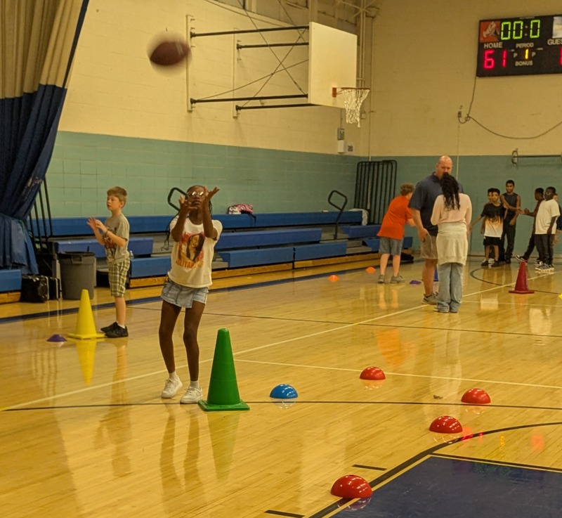 Students are in the gym during Physical Education class practicing throwing footballs. There are several stations established and marked with cones.