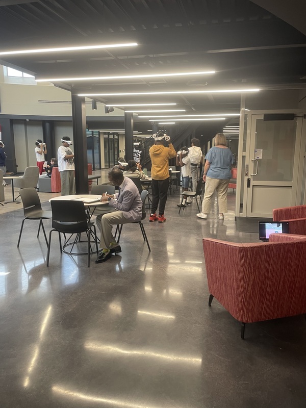 Dr. Saulsberry at table and Jenkins with blue shirt on talking to the students during lesson using VR headsets in library for biology class
