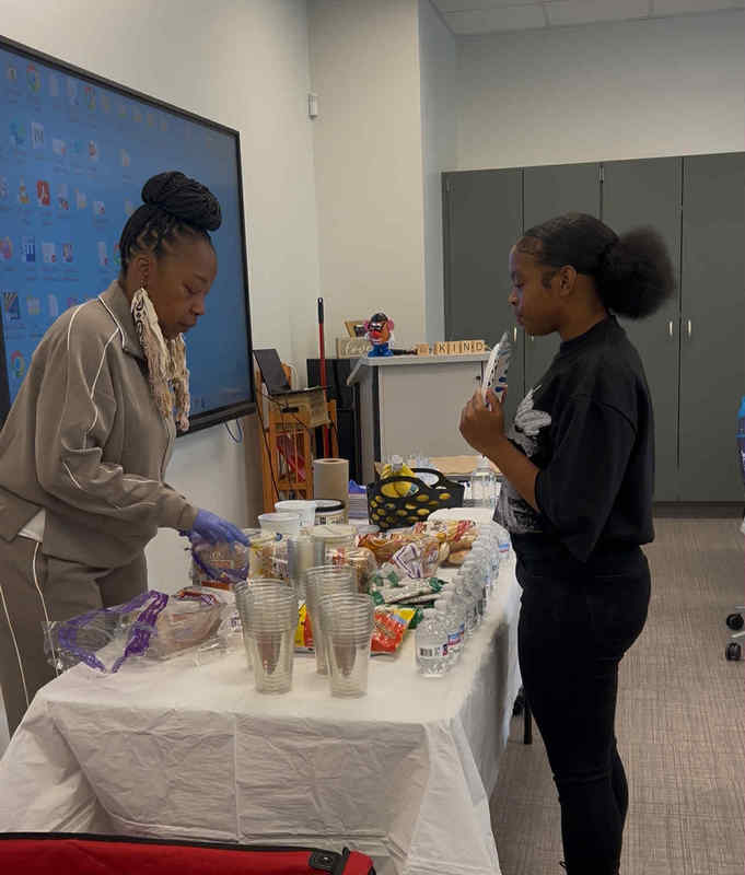 Ms. Johnson, cosmetology teacher, is serving a student a yogurt bowl. There is a table full of various foods for the student to choose from. They are standing in the cosmetology classroom.
