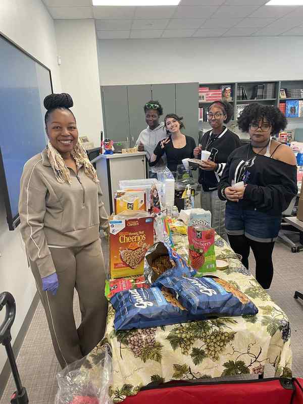 Ms. Johnson posing with four cosmetology students in front of a table of various foods in the cosmetology classroom.