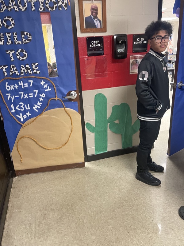 Student standing in front of door  decorated with blue, brown and black writing with green cactuses and pic of principal above