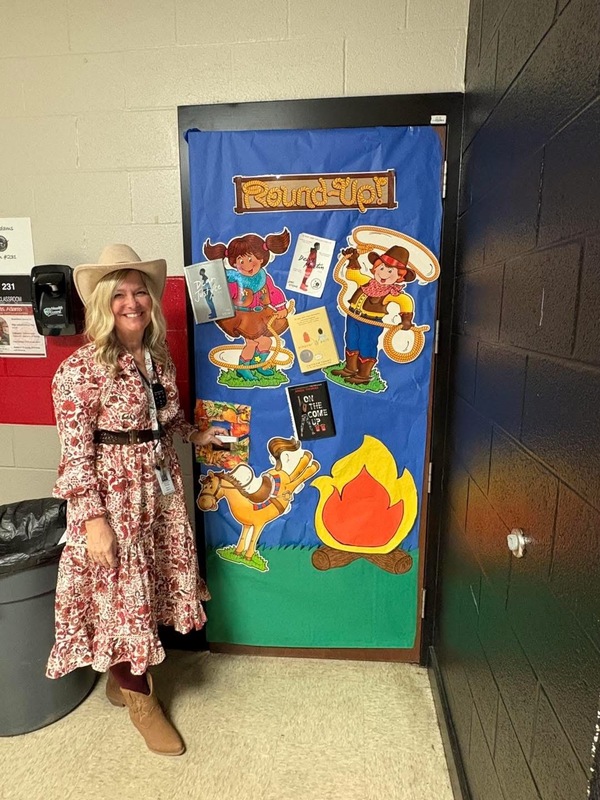 Mrs. Jordan standing in front of her colorful door with blue, yellow, green and orange  with Round Up as title with a cowboy, cowgirl, horse, campfire with books