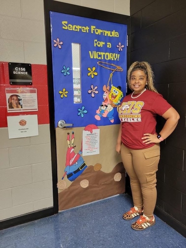 Ms.Kelly posing in front of her decorative door with yellow letters and blue,  brown and red paper with title Secret Formula for a Victory with different colored flowers SpongeBob characters with a lasso