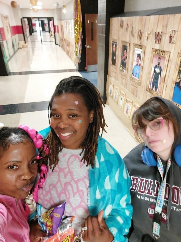 Three teachers  posing for picture dressed like students  with pink hair, one wrapped in blue and white blanket and the third one wearing a grey hoodie with red and white writing 