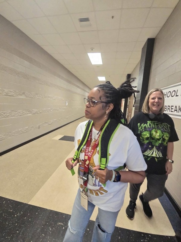 Counselor dressed like student with braid s up in a band, white t-shirt with green necklace and colorful writing on shirt with jeans  on with holes in them as counselor in background with black and green t-shirts laughs in background 