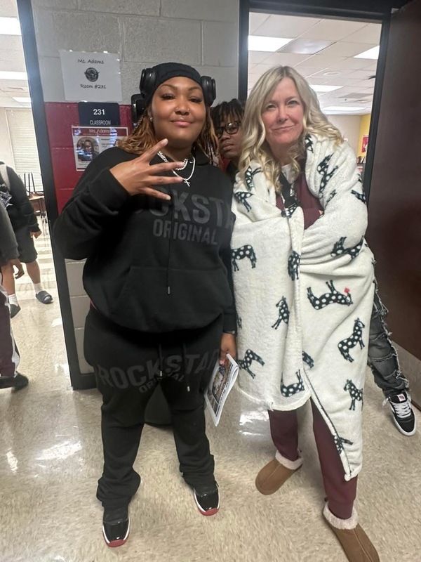 Two teachers - one on the all black, jogging suit and hoodie and the teacher on the right draped in a white and black blanket with brown house shoes on