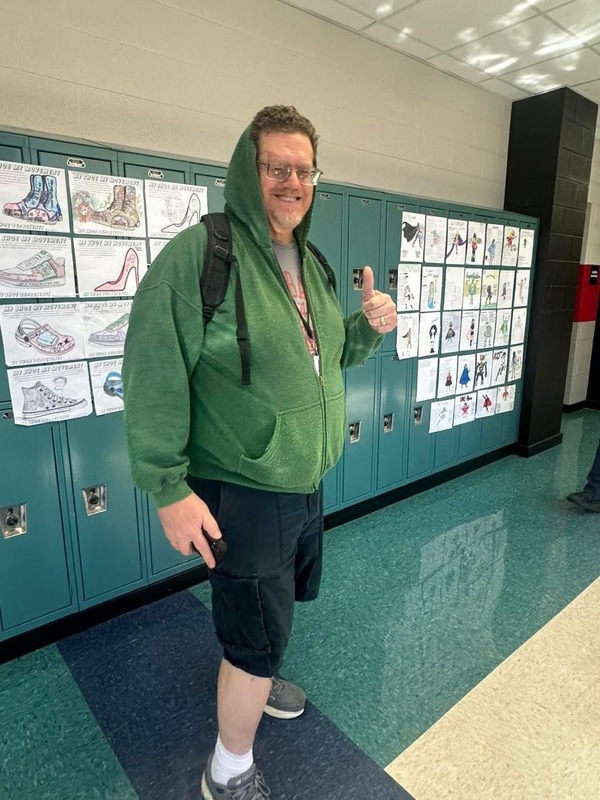 Teacher posing for picture with a green hoodie on with black shorts, white socks, and black tennis shoes in the hallway with lockers behind him and white paper with black writing