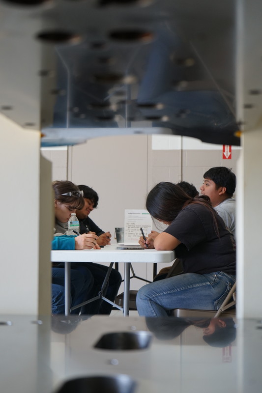 Students sitting at a table writing, the picture is shot through the hole of a machine.