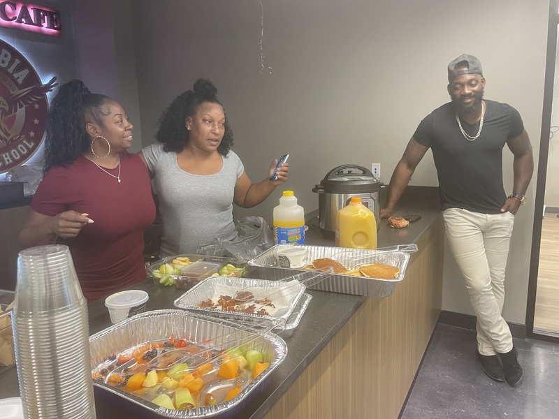 Booster Club president in burgundy, Vice president in gray standing behind food trays and football coach in black shirt and beige pants leaning against the wall