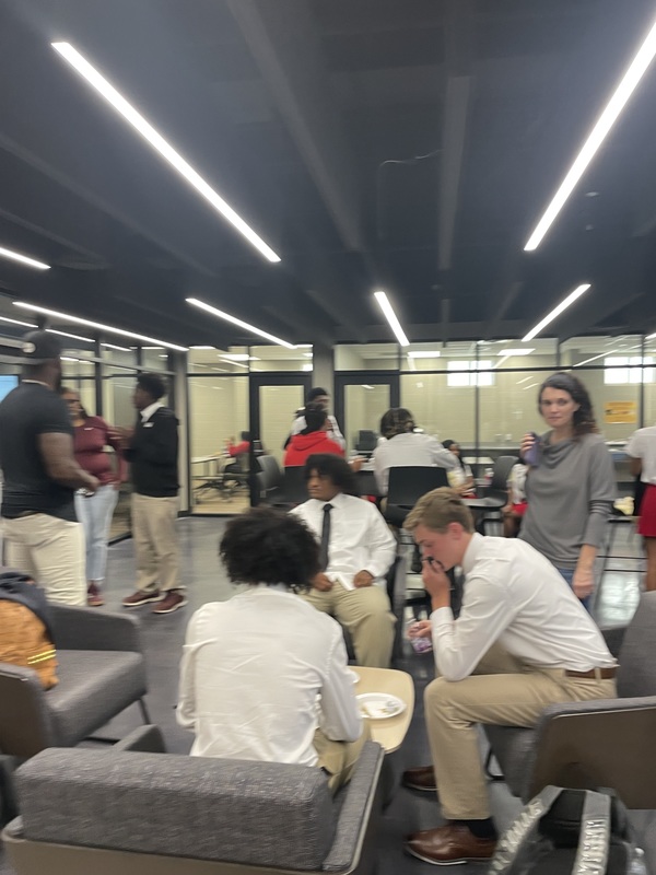 Football players dressed in white shirts and tan pants sitting in gray chairs with a parent standing over them in a gray sweater , the football coachwith back turned wearing black shirt and pants and other students in the background
