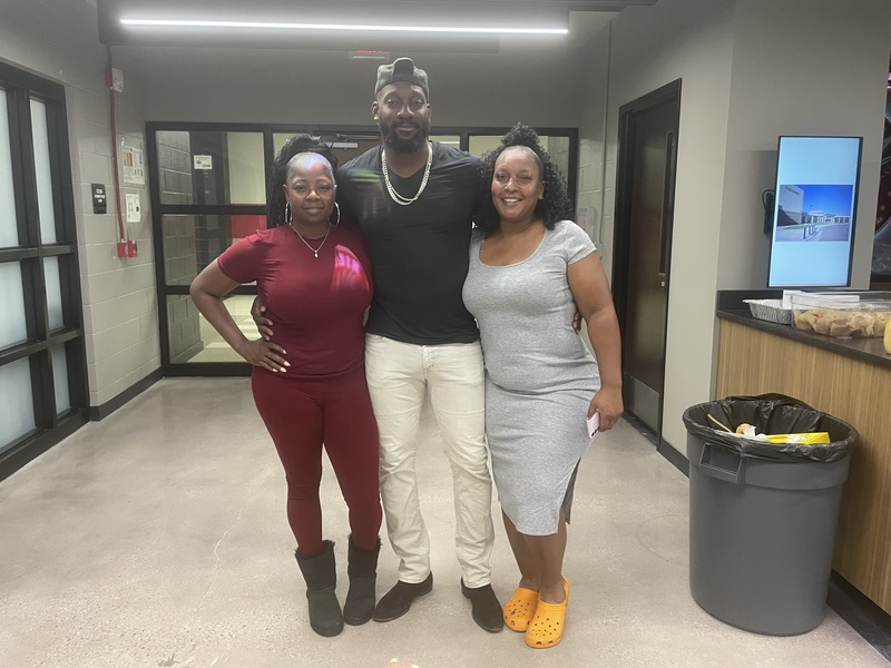 Booster Club president dressed in burgundy shirt and pants, football coach in mddle with black shirt and tan pants and vice president in light gray dress and yellow shoes