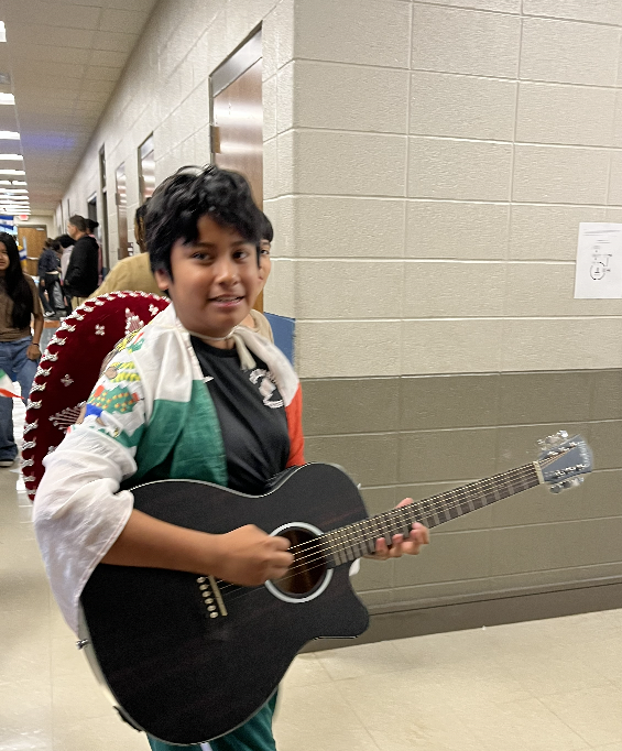 A student joins the parade wearing a Mexican flag draped around his shoulders,  a sombrero hanging down his back and carrying a guitar