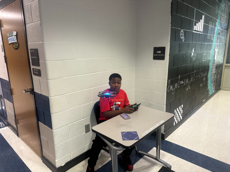 A student in a red shirt at a desk displays a drone from the drone club while holding its remote control.