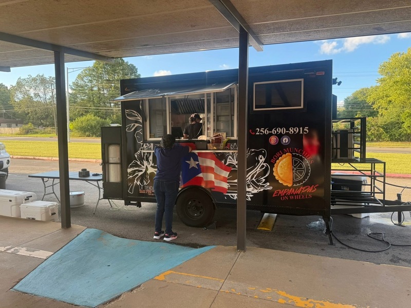 A woman stands next to a food truck, which features a bright design and a menu showcasing various meal options.