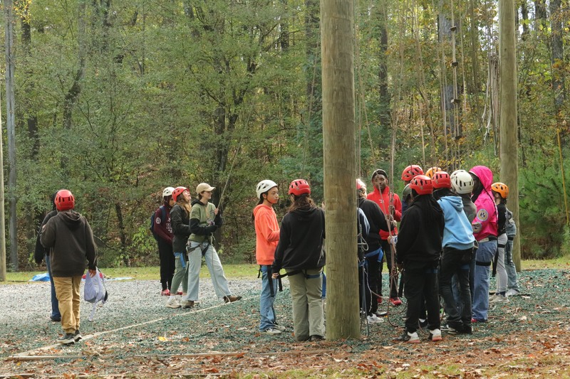 A group of scholars gathered around a rope course, discussing their next challenge in an outdoor adventure setting.