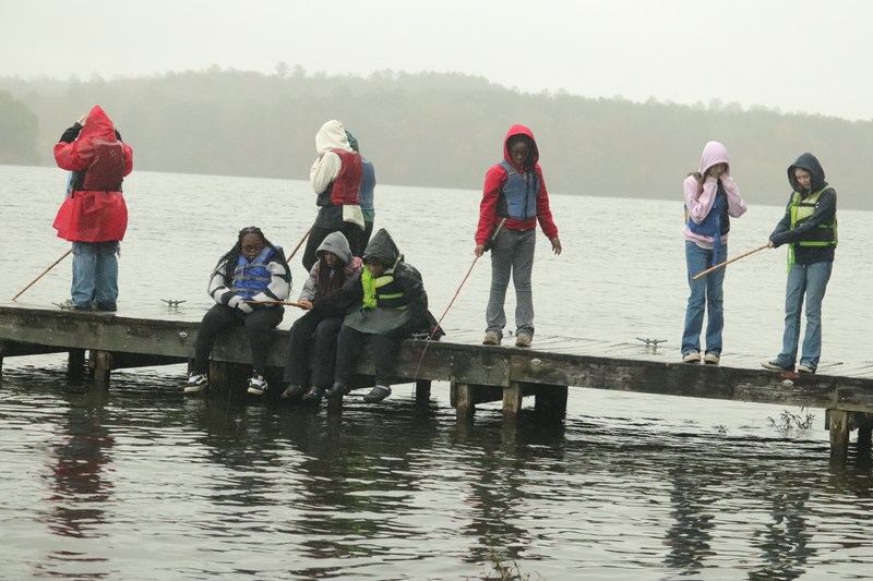 Several scholars gathered on a dock, fishing and enjoying the view of the water and surrounding scenery on a cloudy day.