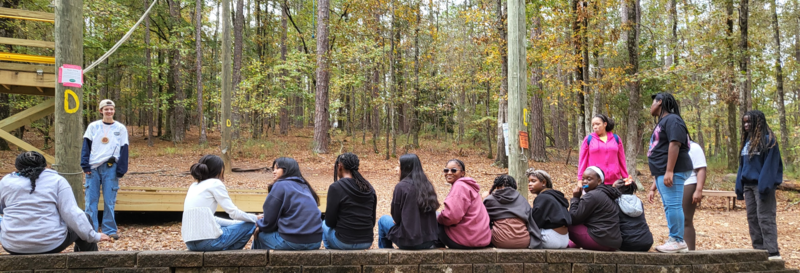 A group of scholars sitting on a wooden bench surrounded by trees in a serene forest setting.