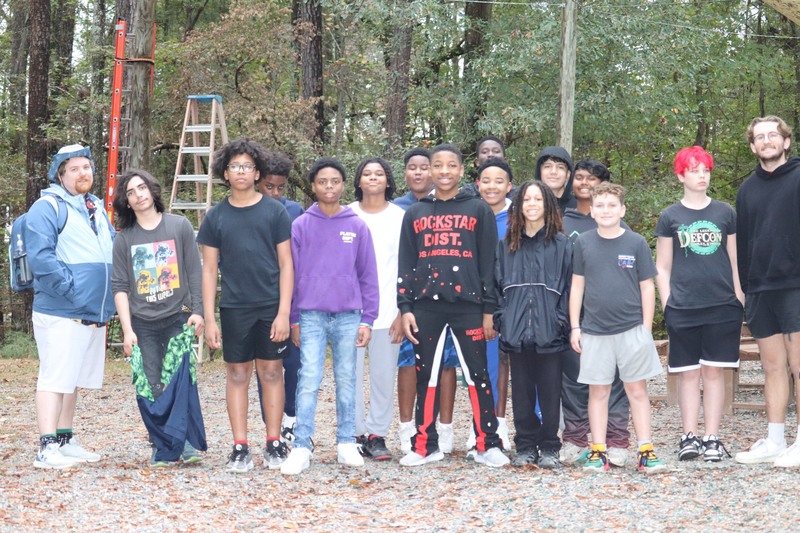 A group of young people stands together in front of a large tree, smiling and enjoying each other's company.