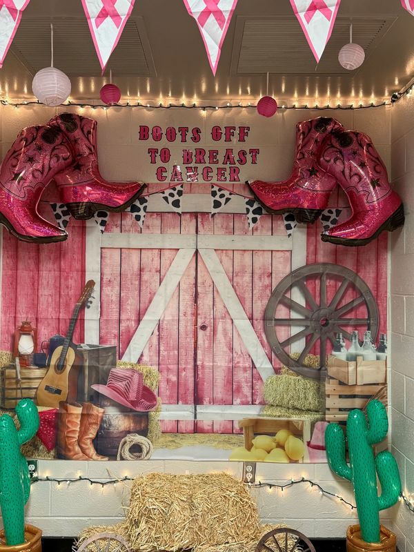 Boots Off to Breast Cancer Wall with 2 big, pink boots on each side of a mural depicting a pink and white barn with green cactuses on each side and a guitar boots pink hat bucket on left side with a hay stacj in front