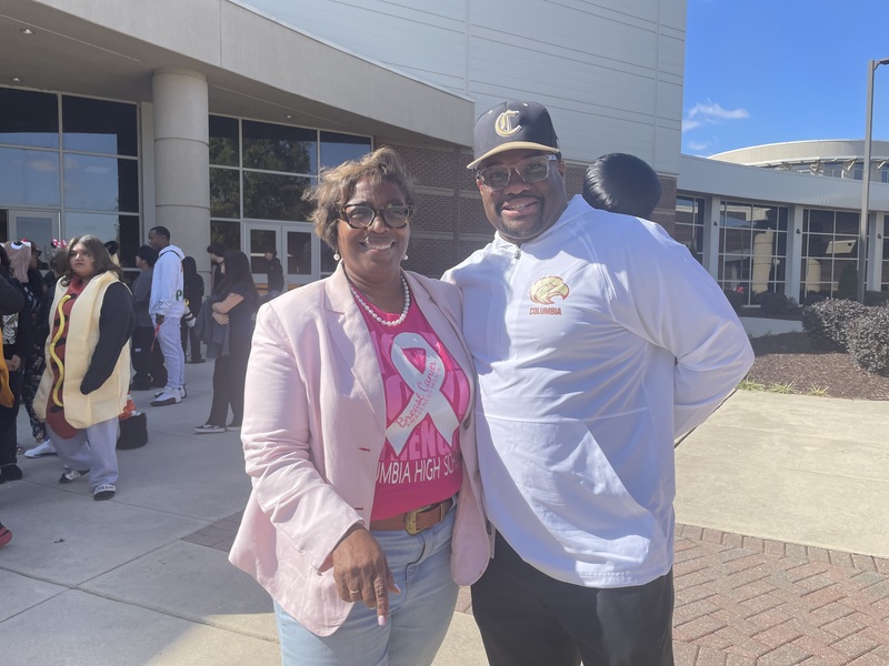 FBLA Sponsor on left wearing a light pink jack and hot pink shirt with anding with principal who is wearing a white shirt, dark pant and dark blue and gold hat with a C on it in front of school and students in the background