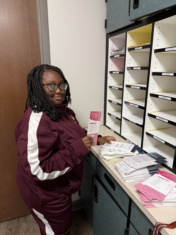 Student standing at boxes in the front office wearing a marroon and white jogging suit passing out breast cancer awareness panplets
