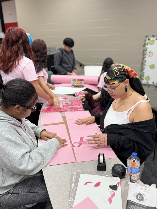 Six students from FBLA sitting at table with pink paper, ribbons, glue, tape making Breast Cancer Awareness signs