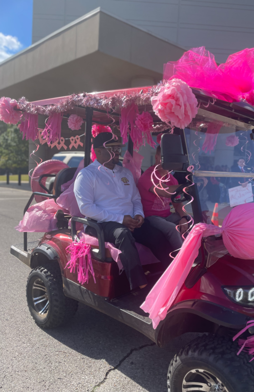 Principal Lowery in a pink shirt and principal with white shirt and dark colored baseball cap sittting a golf cart that is decorated in pink paper and balloons for Breast Cancer