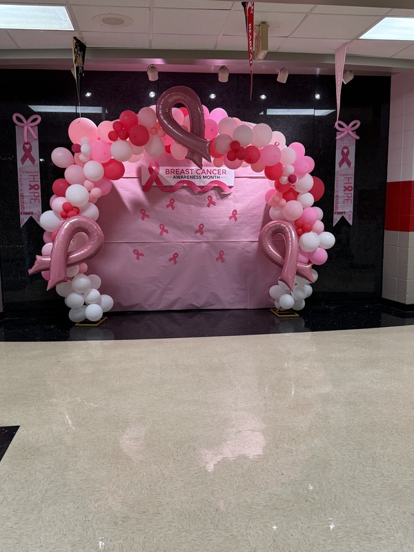 Pink and white balloon arch with three pink ribbons around arch with pink bulletin board paper in the middle with pink bows and breast cancer sign at the top
