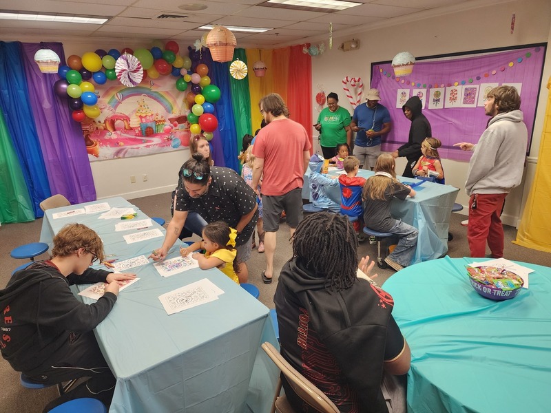 Esports students and parents at tables with students helping them color on tables with blue cloths wall has green, blue, purple curtains on each side with colorful balloons in the middle