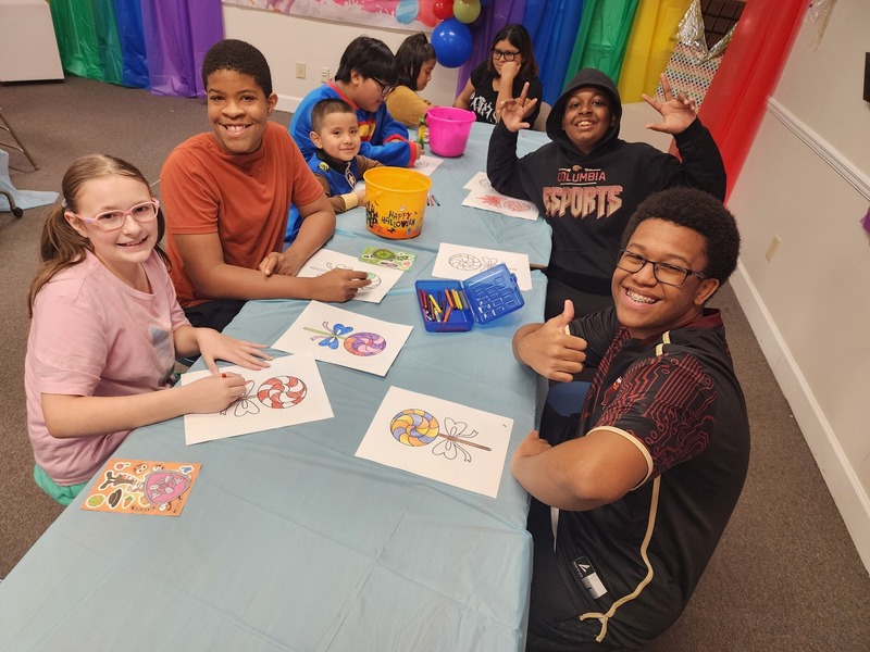 Students and 2 esports members at table with blue cloth and white coloing sheet and a blue container of markers and yellow and pink pails
