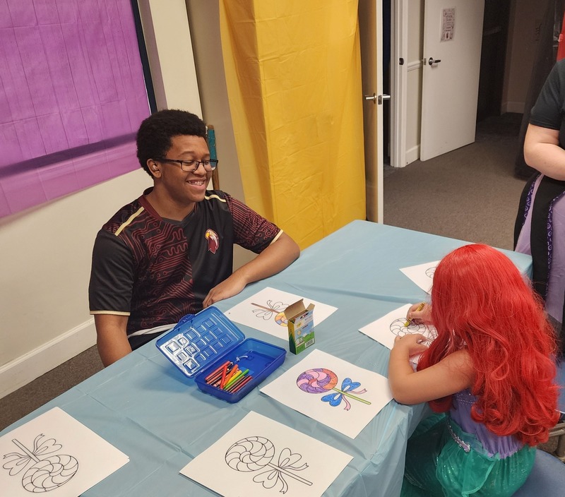 Esports member sitting at table with light blue cloth with marron and black shirt smiling sitting across from student with long, red hair while she is coloring a color sheet with a blue open container of colored pens and a crayon box in front of her