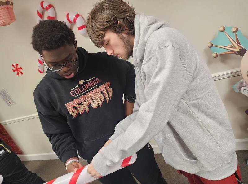 2 esports team members witht the one on the right wearing a black and red esports shirt and the other in a light gray sweatshirt hoodie working together putting red stripe on a white pole