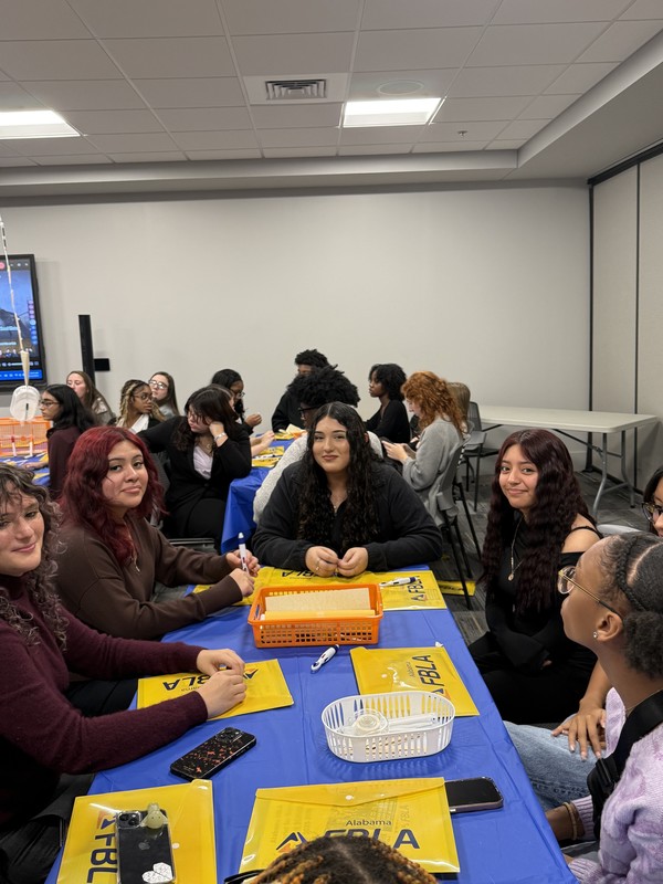 Students in FBLA sitting around table with blue cloth and yellow folders with FBLA on it with a white container on it