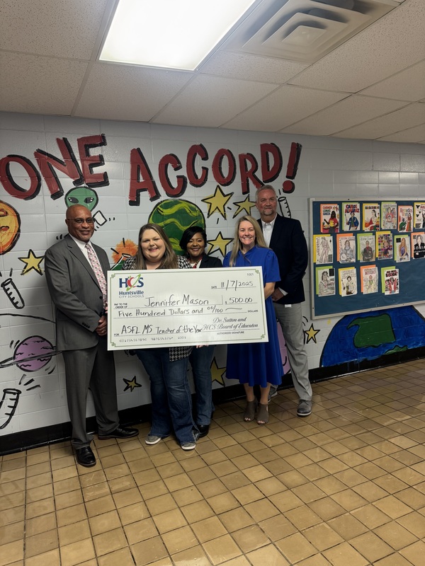Collaborative teacher, Principal Shoulders, and District Representatives stands in front of a wall displaying a large check of $500, smiling and posing for the photo.
