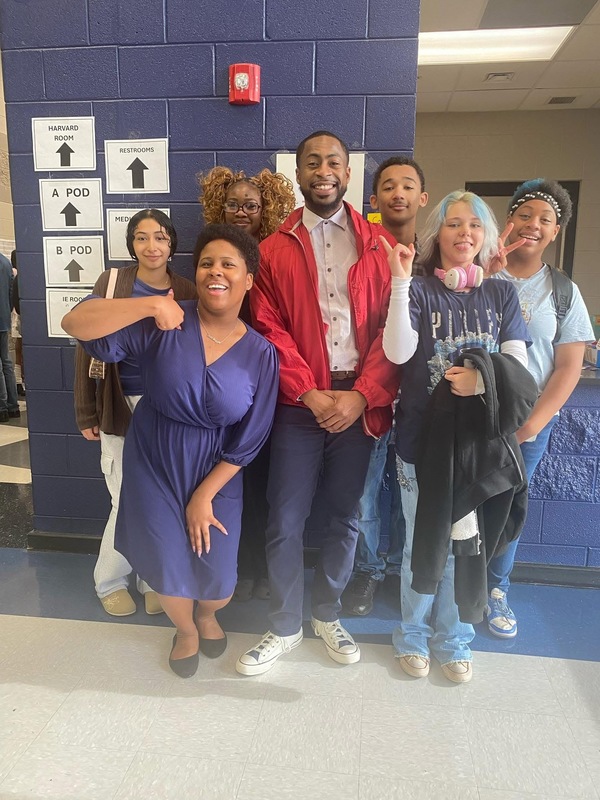 7 theatre students with teacher in middle wearing red jacket, white shirt, dark blue pants and tennis shoes posing for pic in front of caferia with dark blue wall, red fire part and white and black signs
