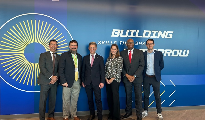 Dustin Daehn, Zac McWhorter, Dr. Mackey, Carolyn Wade, Dr. Sutton, and Craig Williams pose in front of a wall in the Huntsville Center for Technology that says Building Skills that Shape Tomorrow