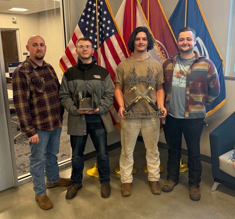 Welding students pose with their welding instructors in front of three flags