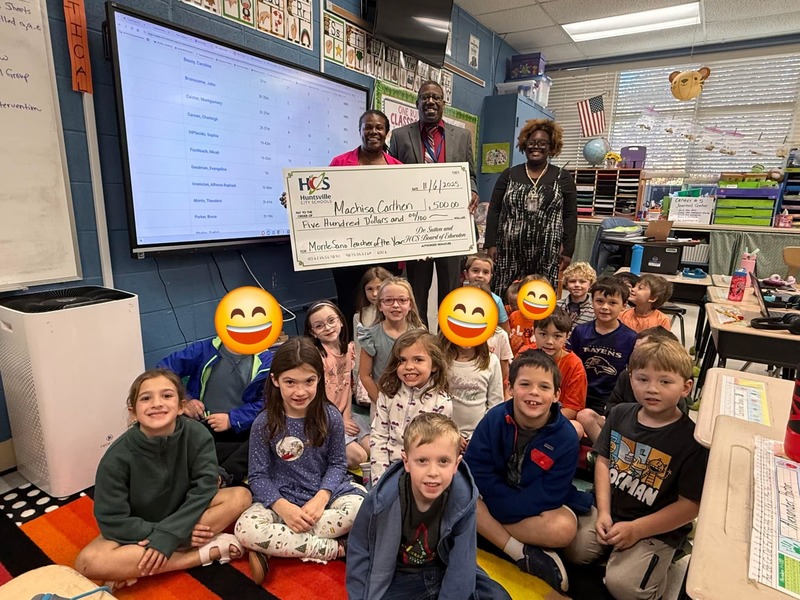 students sitting on carpet with teacher and principal standing behind them holding large check for $500