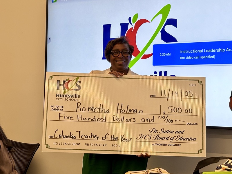 Mrs. Holman standing in front of white board with HCS logo holding a big check written out to her for $500 for Teacher of the year