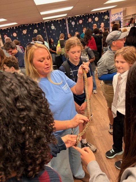 Lady in a blue shirt presents a small patterned snake to a group of interested students in a crowded classroom setting.