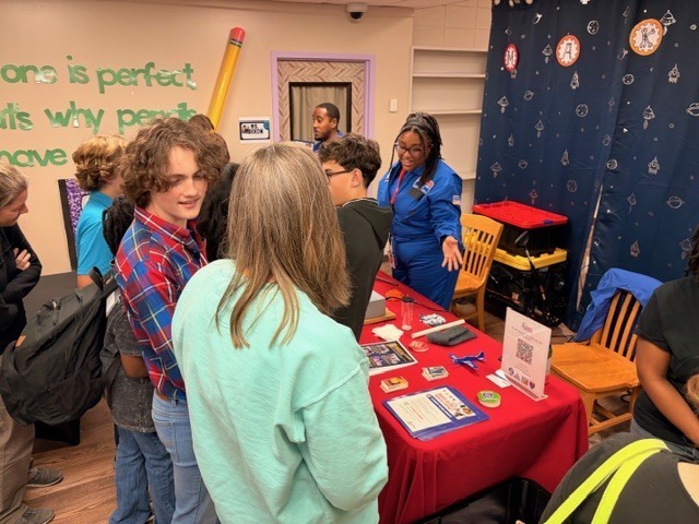 A group of diverse students gathers around a red-clothed table where a woman in a blue flight suit explains educational materials, next to a space-themed curtain