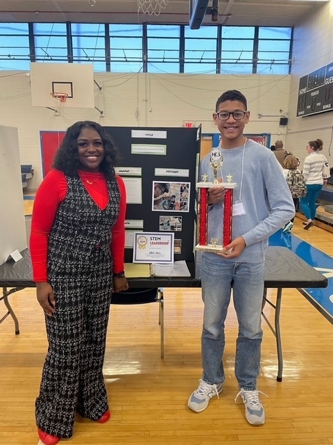 ASFL science teacher in a red top and tweed jumpsuit stands next to an ASFL student holding a STEM leadership trophy in a school gymnasium setting.