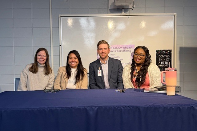 Four smiling professionals sit behind a table draped in a navy blue cloth in front of a whiteboard displaying STEAM-related text for our STEM panel