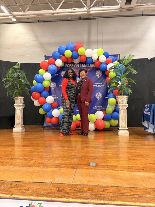 ASFL science teacher and principal stand smiling on a wooden stage in front of a red, blue, green, and white balloon arch celebrating HCS Middle School District Science Fair