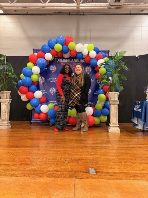 ASFL Science and Collaborative teacher stand smiling on a wooden stage in front of a red, blue, green, and white balloon arch celebrating HCS Middle District Science Fair