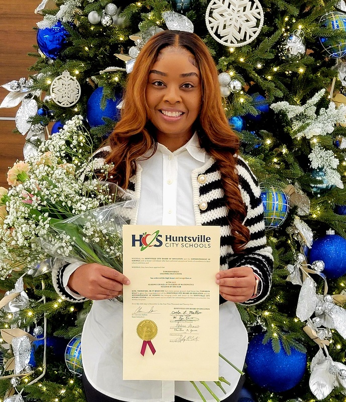 Ms. Kelly standing in front of decorated Christmas tree holding an HCS award